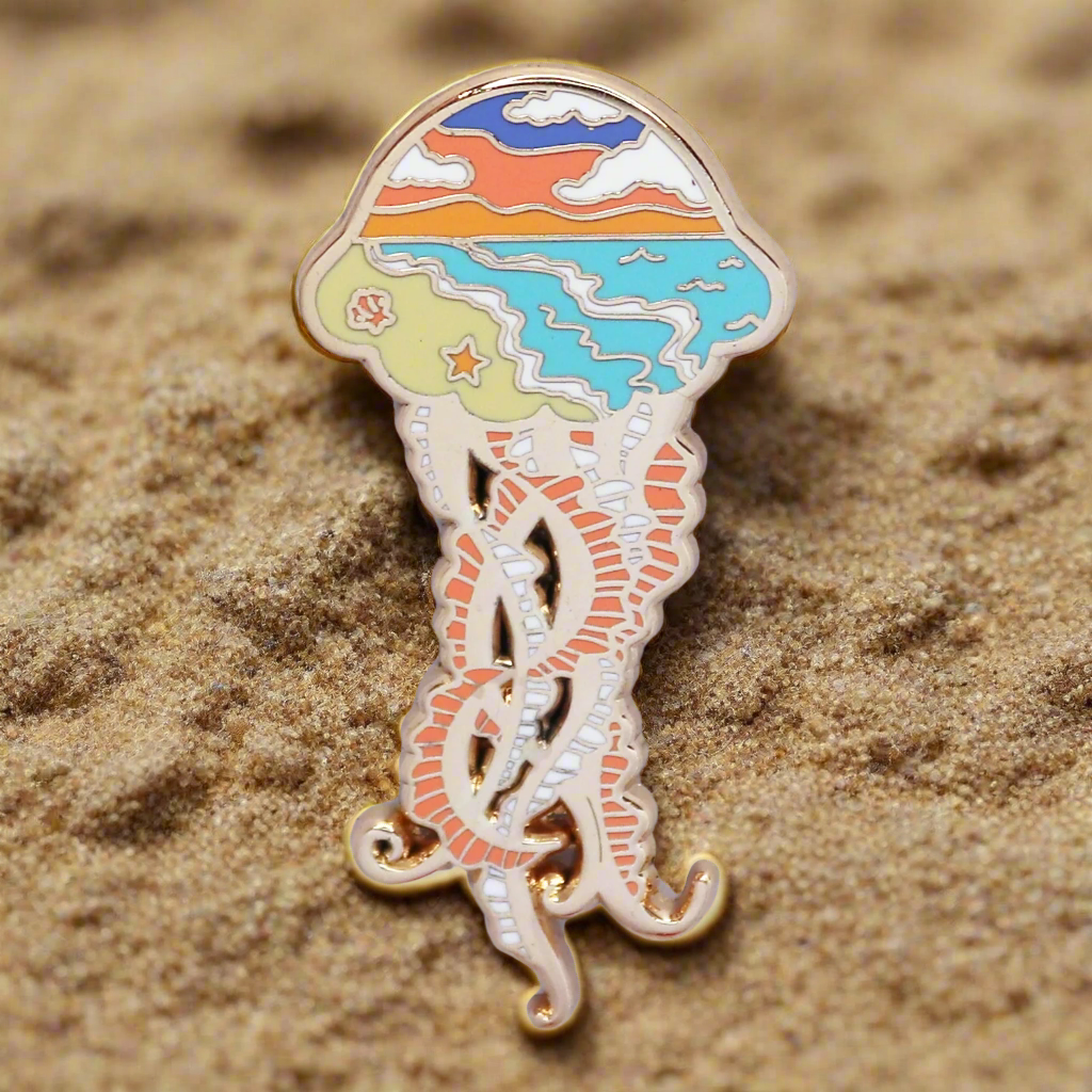 A gold metal enamel pin shaped like a jellyfish with a beach and ocean silhouette design, placed on a dual-tone background.