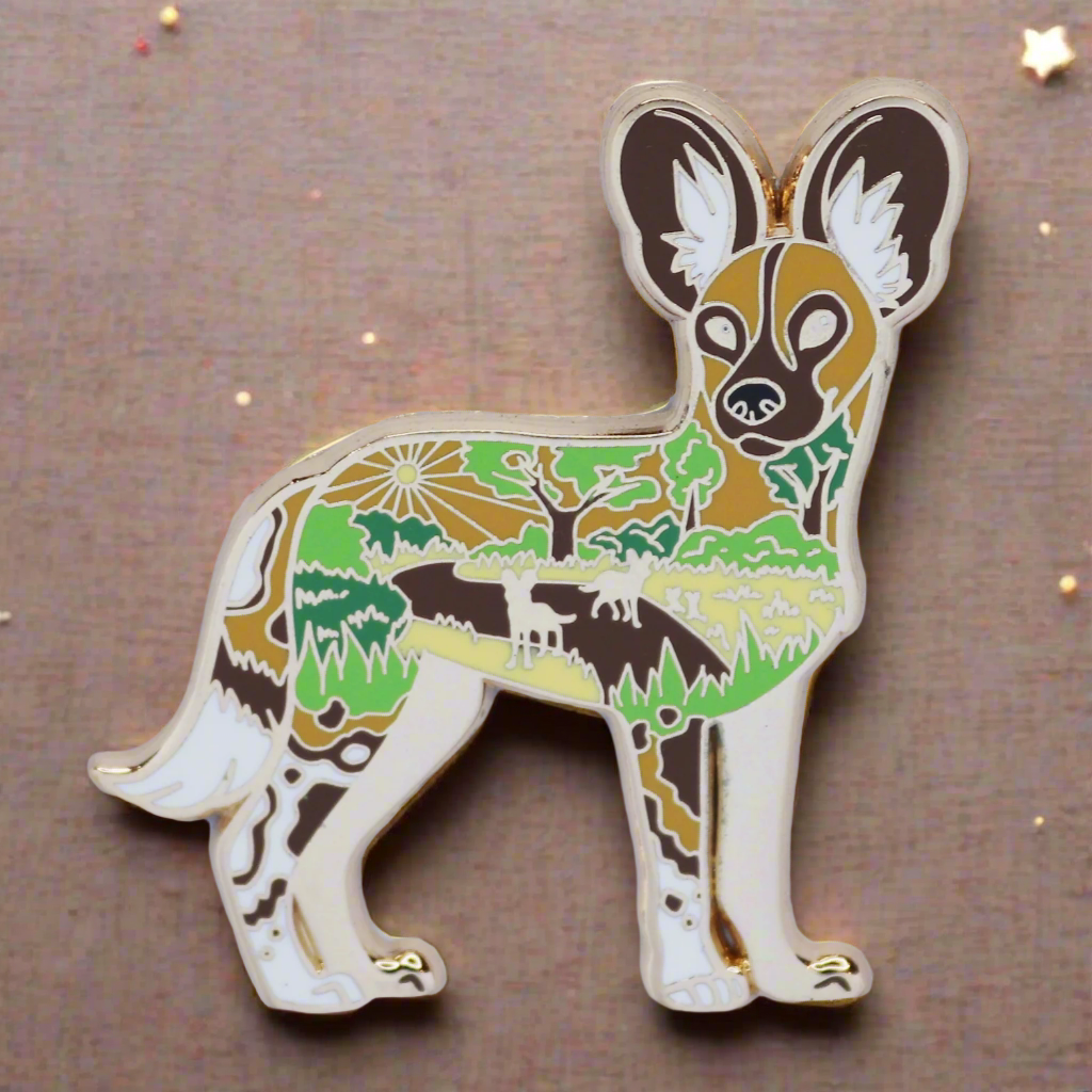 A gold metal enamel pin shaped like an African Wild Dog with a landscape background.