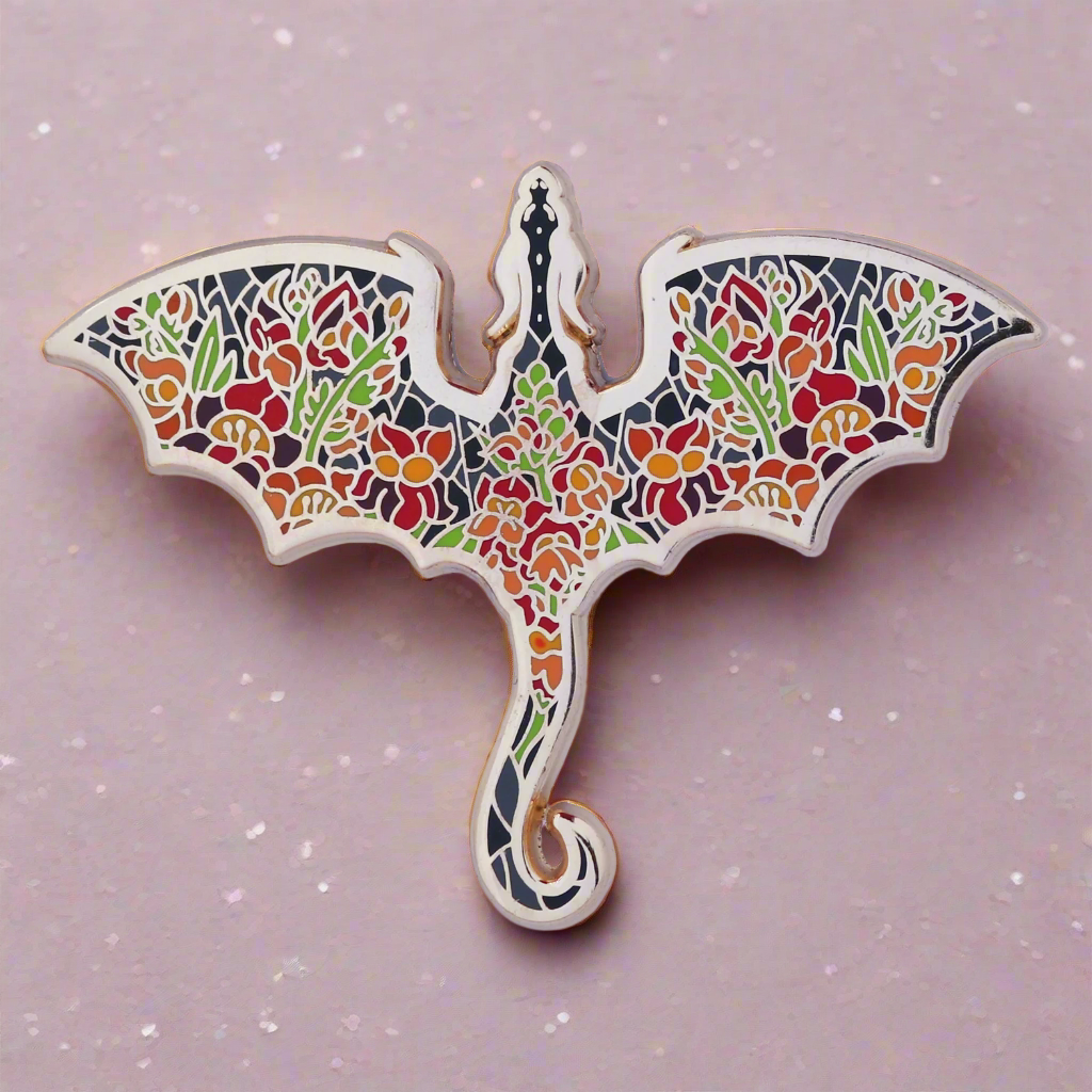 An enamel pin with a dragon silhouette design, featuring floral patterns in various colors on a gold metal background.