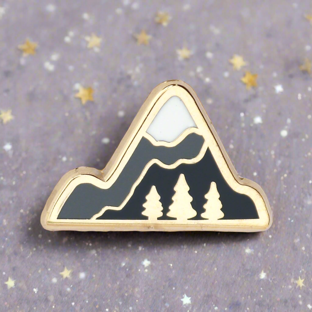 A gold metal framed enamel pin shaped like a mountain range with trees.