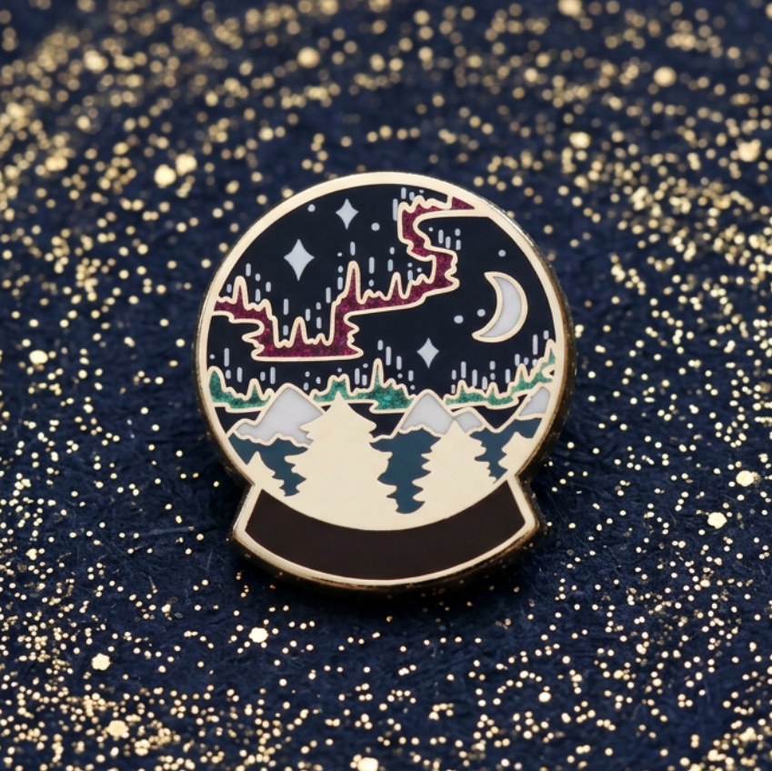 A circular enamel pin shaped like a snow globe, featuring a night sky with stars and a crescent moon above snowy mountains and trees, outlined in gold metal on a light background.