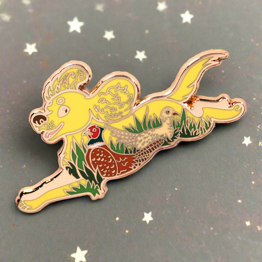 A gold metal enamel pin featuring a spaniel dog with a pheasant in the background, on a two-tone background with pink at the top and yellow at the bottom.