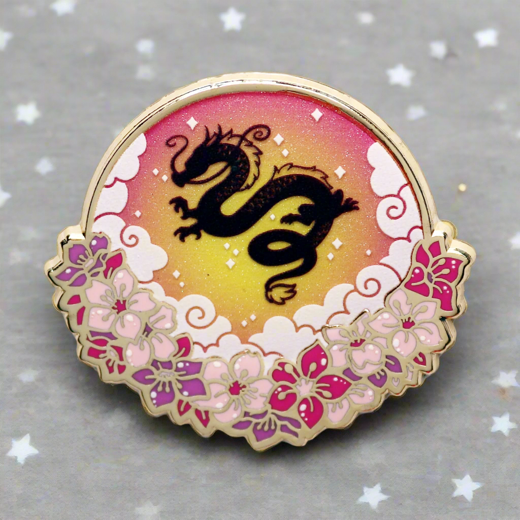 An enamel pin with a gold metal border featuring a silhouette of a dragon against a gradient background with pink and purple hues, and floral patterns.
