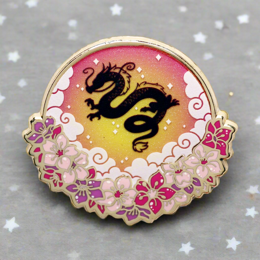 An enamel pin with a gold metal border featuring a silhouette of a dragon against a gradient background with pink and purple hues, and floral patterns.