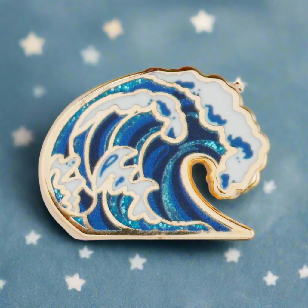 A gold metal, hard enamel pin with a design of a blue wave with glitter details.